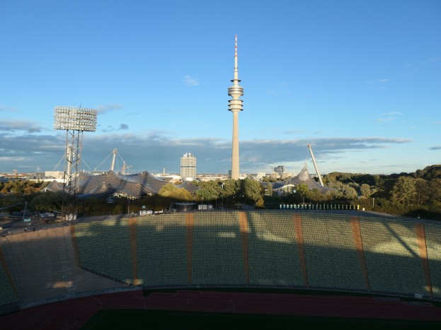 Olympiastadion in Berlin, Deutschland, mit dem Fernsehturm im wolkenverhangenen Hintergrund, umgeben von Bäumen, Gebäuden und beleuchteten Strukturen.