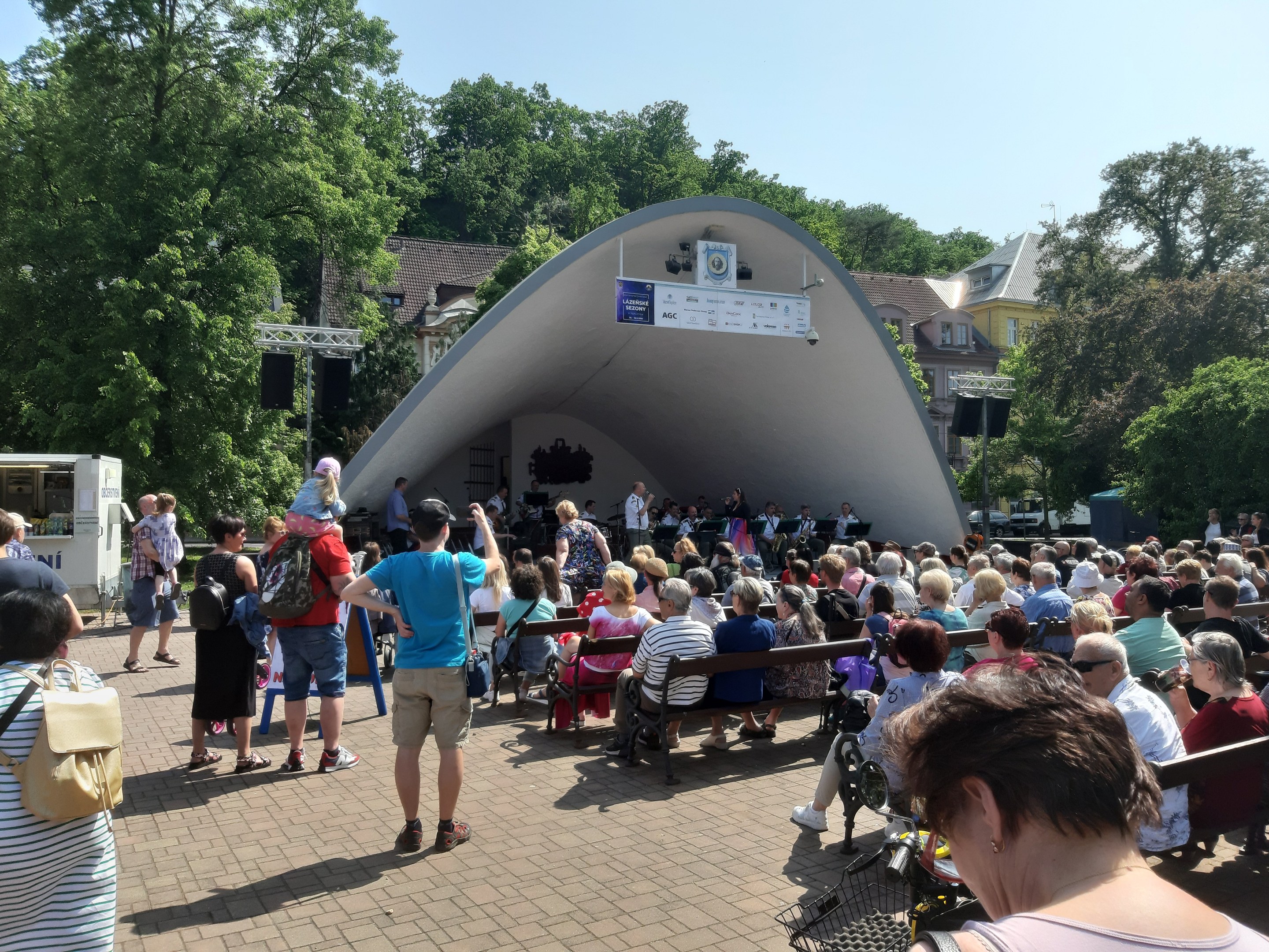 Menge Menschen auf Bänken und Stehplätzen vor einer Bühne mit einer Kamera, einem Mikrofonständer, einer Fahne, einem Metallrahmen, Bäumen, Gebäuden und einem bewölkten Himmel bei einem Sommerkonzert im Park.
