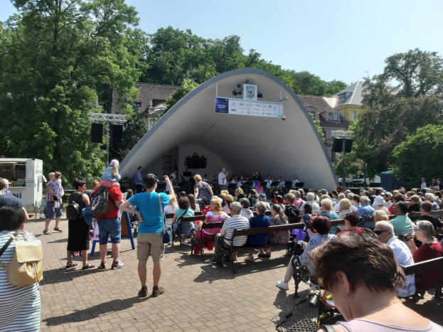 Menge Menschen auf Bänken und Stehplätzen vor einer Bühne mit einer Kamera, einem Mikrofonständer, einer Fahne, einem Metallrahmen, Bäumen, Gebäuden und einem bewölkten Himmel bei einem Sommerkonzert im Park.
