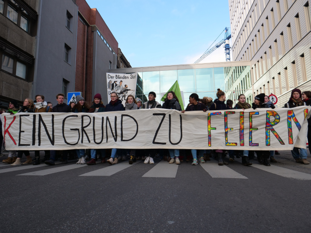 Demonstranten mit einem Banner "Kein Grund zu Feiern" gegen deutsche Sparmaßnahmen, mit Gebäuden, einem Kran und einem bewölkten Himmel im Hintergrund.
