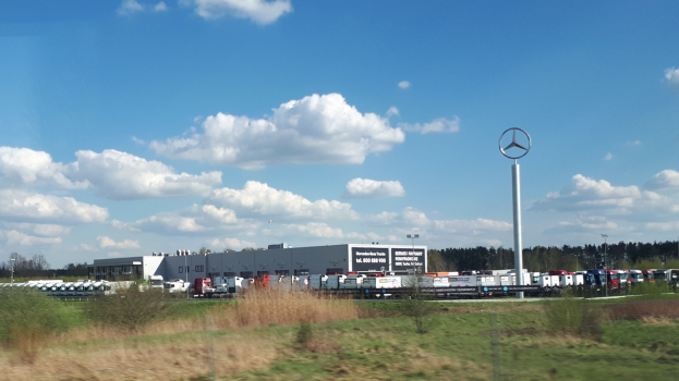 Ein Mercedes-Benz-Werk in Deutschland mit Fahrzeugen auf der Straße, umgeben von Grünflächen und Gebäuden unter einem bewölkten Himmel.