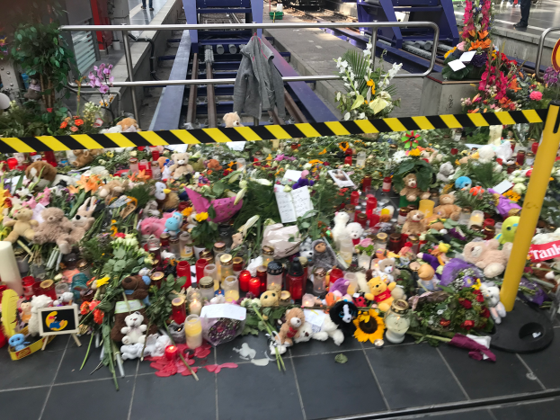 Ein Bahnsteig in London, England, geschmückt mit Teddybären, Blumensträußen, Kerzen und anderen Gegenständen als Erinnerung an eine Tragödie, mit Menschen im Hintergrund.
