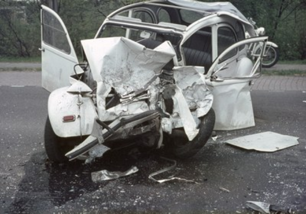Ein schwerbeschädigtes Auto steht mit Beschädigungen an der Front auf dem Seitenstreifen, mit Bäumen, einem Gebäude und einem anderen Fahrzeug im Hintergrund.