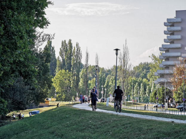 Eine Gruppe von Menschen, die auf Fahrrädern einen Parkweg entlangfahren, mit Gras, Bäumen, Straßeninfrastruktur, Kraftfahrzeugen und Gebäuden im Hintergrund unter einem bewölkten Himmel.