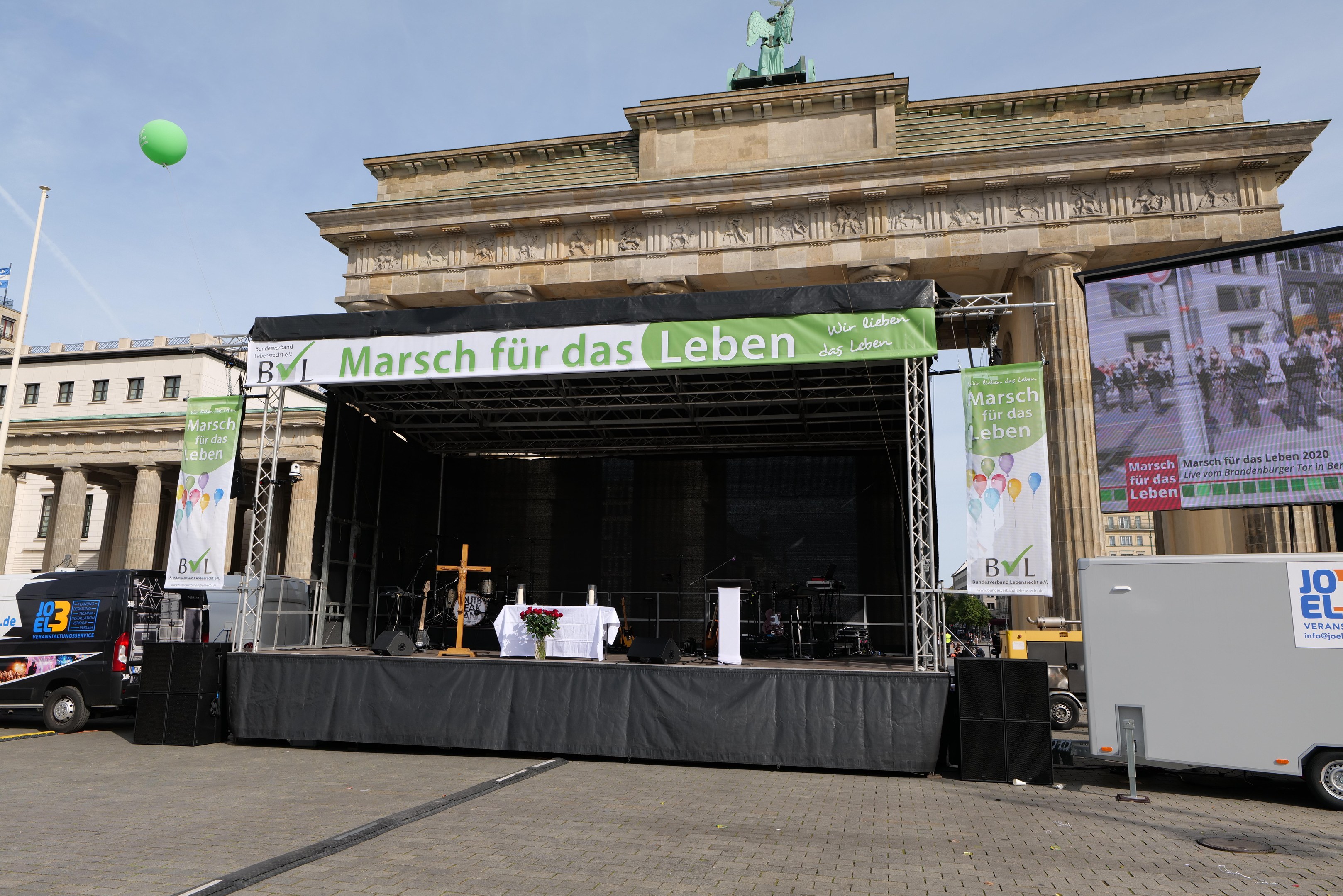 Eine Bühne vor dem Brandenburger Tor in Berlin, Deutschland, mit einem Tisch mit einer Blumenvase, einem Kreuzsymbol, Transparenten mit Text, Lautsprechern und anderen Gegenständen, sowie Fahrzeugen auf der Straße und Gebäuden im Hintergrund.