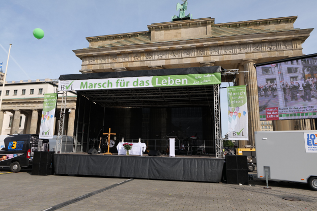Eine Bühne vor dem Brandenburger Tor in Berlin, Deutschland, mit einem Tisch mit einer Blumenvase, einem Kreuzsymbol, Transparenten mit Text, Lautsprechern und anderen Gegenständen, sowie Fahrzeugen auf der Straße und Gebäuden im Hintergrund.