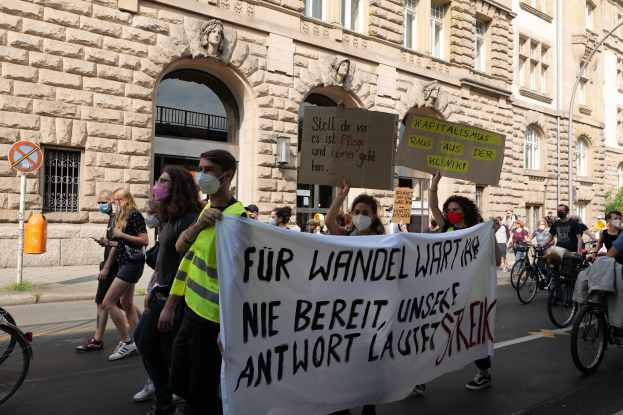 Eine Gruppe von Demonstranten marschiert eine Straße entlang, einige halten Schilder und andere fahren Fahrräder, mit einem historischen Gebäude im Hintergrund mitbogigen Fenstern, Säulen und Skulpturen.