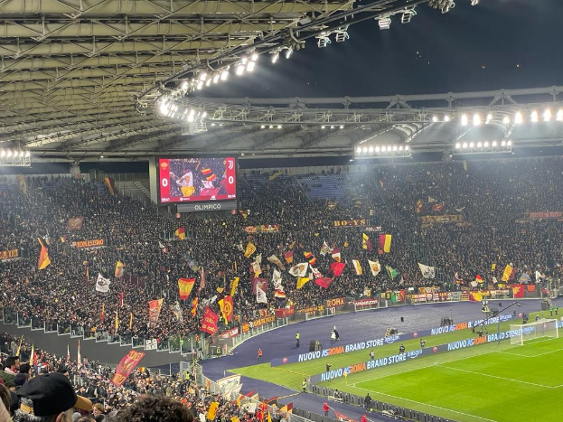 Eine große Menschenmenge in einem Stadion schwenkt Fahnen, mit Bannern, Gras, einem Netz und einer beleuchteten Decke, in Erwartung eines Fußballspiels.
