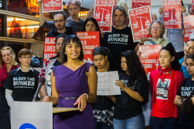Eine Black Lives Matter-Aktivistin spricht an einem Podium mit Mikrofon vor einer Menge mit Schildern, vor Lichtern und anderen Gegenständen.
