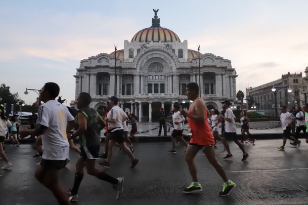Eine Gruppe von Menschen, die bei einem Marathon vor einem großen Gebäude laufen, mit Straßenschildern, Straßenlaternen, Bäumen und einem bewölkten Himmel im Hintergrund.