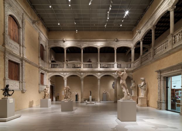 Großer Raum in einem Museum mit zahlreichen Statuen und Skulpturen auf Sockeln, architektonischen Elementen wie Säulen und Geländern und einigen Besuchern unter Deckenbeleuchtung.