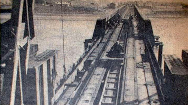 Ein Schwarz-Weiß-Foto einer Bahntrasse mit einer im Hintergrund befindlichen Brücke, umgeben von Werkzeugen und Materialien.