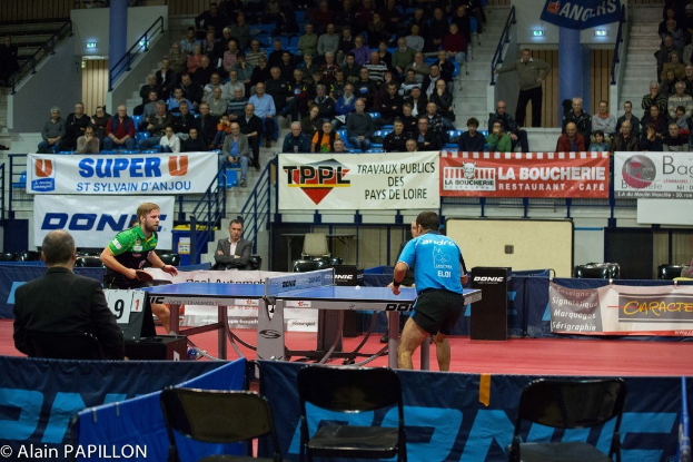 Indoor-Tischtennis-Match mit zwei Spielern, mehreren stehenden Zuschauern, sitzendem Publikum, leeren Stühlen und Werbeplakaten im Hintergrund.