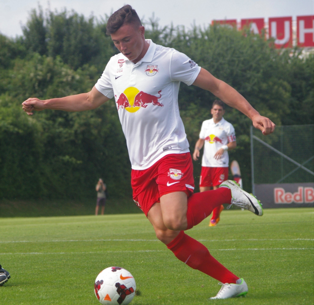 Ein professioneller Fußballspieler kickt einen Ball auf einem grünen Feld mit Bäumen und einem klaren blauen Himmel im Hintergrund, wobei eine Tafel mit der Aufschrift "RB Leipzig" sichtbar ist.