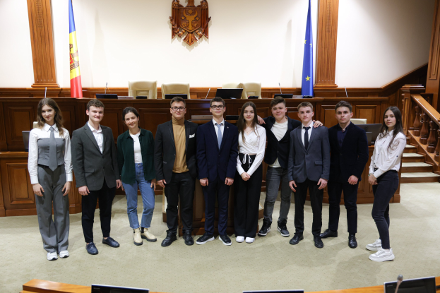 Gruppe von strahlenden Menschen, die vor einem Gerichtssaal mit Monitoren und Mikrofonen im Vordergrund und einer hölzernen Plattform mit Fahnen und Stühlen im Hintergrund posieren.