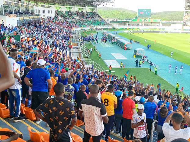 Eine große Menschenmenge versammelt sich vor einem eingezäunten Fußballfeld, wobei einige auf Stühlen sitzen und andere stehen, während Hügel und ein klarer blauer Himmel die Kulisse bilden während der 2019 African Cup of Nations.