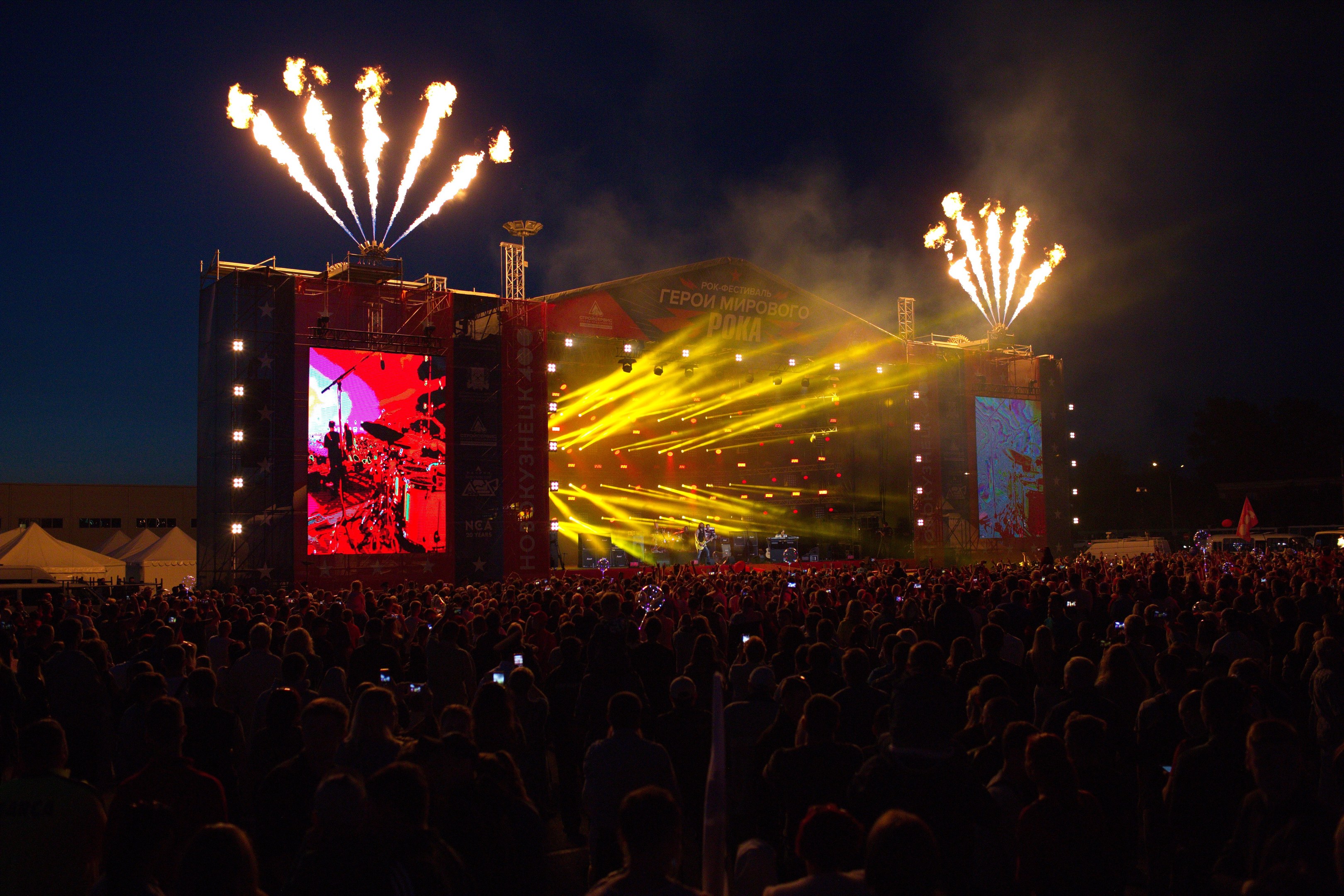 Große Menschenmenge auf einem Musikfestival schaut auf einer hell erleuchteten Bühne mit Bannern, umgeben von Zelten und Feuerwerk am Nachthimmel.