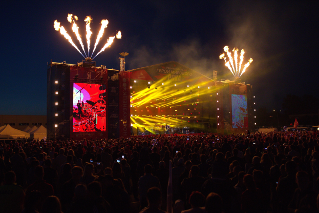 Große Menschenmenge auf einem Musikfestival schaut auf einer hell erleuchteten Bühne mit Bannern, umgeben von Zelten und Feuerwerk am Nachthimmel.