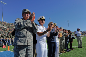 Eine Gruppe von Menschen steht auf einem Fußballfeld und klatscht, mit Zuschauern auf den Rängen, Laternenmasten und einem klaren blauen Himmel im Hintergrund.