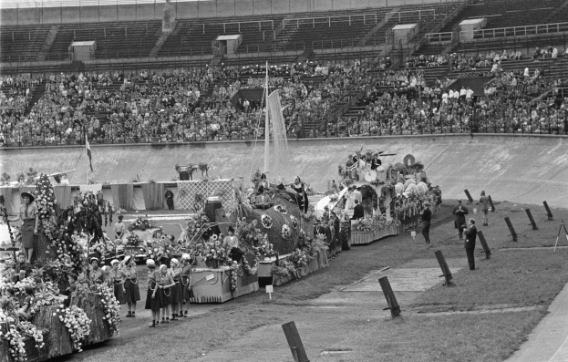 Schwarzes und weißes Foto eines Umzugs in einem Stadion mit Menschen, die stehen und sitzen, einem zentralen Springbrunnen und Blumenbouquets auf den Fahrzeugen.
