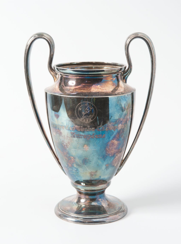 Der UEFA-Champions-League-Pokal, ein stählerner Becher mit zwei Griffen und einem blauen und silbernen Farbschema, der mit "UEFA Champions League" auf einem weißen Hintergrund eingraviert ist.