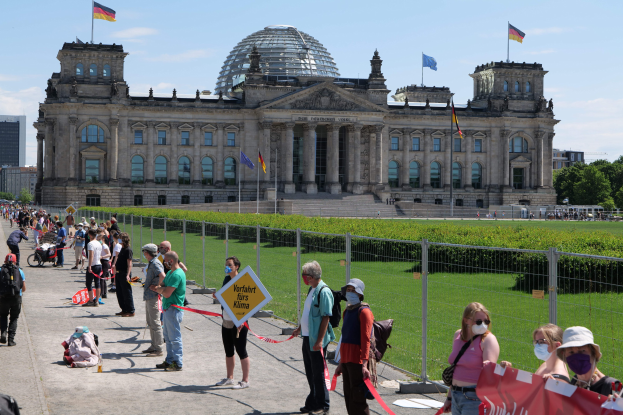 Eine Gruppe von Menschen steht vor dem Reichstaggebäude in Berlin, einige tragen Mützen und Schutzbrillen, andere halten Schilder und einen Kinderwagen, mit Geländern, Pflanzen, Gebäuden, Flaggen und Bäumen im Hintergrund.