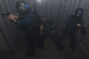 Eine Gruppe von Polizisten in Einsatzausrüstung, einer hölt eine Waffe, steht in der Nähe eines Metallbehälters und einer Leiter in einem beengten Raum.