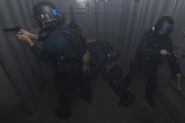 Eine Gruppe von Polizisten in Einsatzausrüstung, einer hölt eine Waffe, steht in der Nähe eines Metallbehälters und einer Leiter in einem beengten Raum.