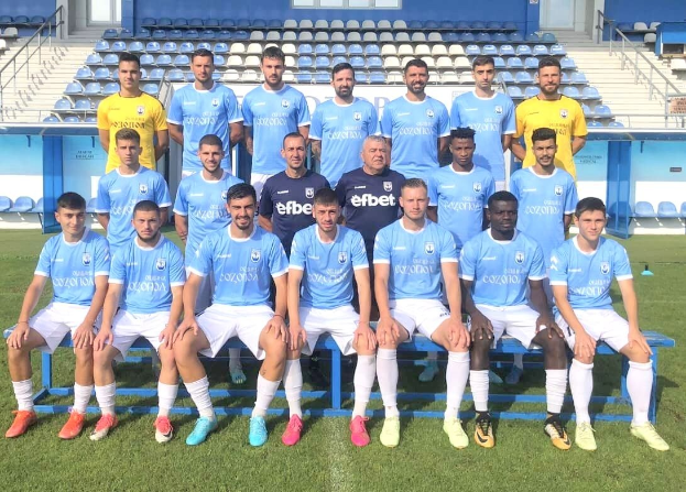 Eine Gruppe von Männern, einige sitzen auf Bänken und andere stehen auf einem Fußballfeld, mit Tribünen und architektonischen Elementen im Hintergrund, wahrscheinlich feiern sie einen Mannschaftssieg.