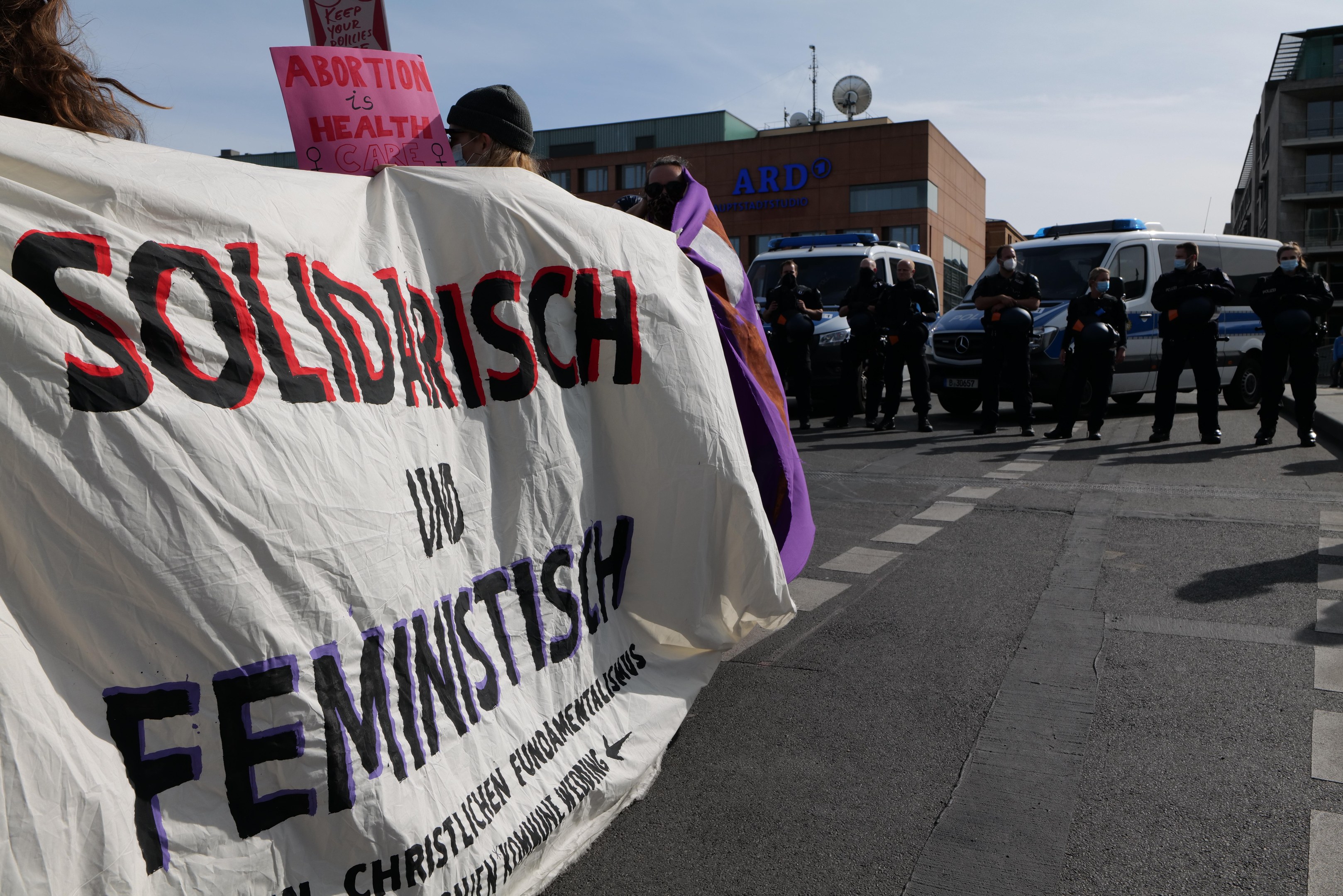 Eine Gruppe von Menschen marschiert die Straße entlang, mit einer Fahne mit der Aufschrift "Solidarität und Feminismus", mit parkenden Fahrzeugen am Rand und Gebäuden im Hintergrund unter einem klaren blauen Himmel.