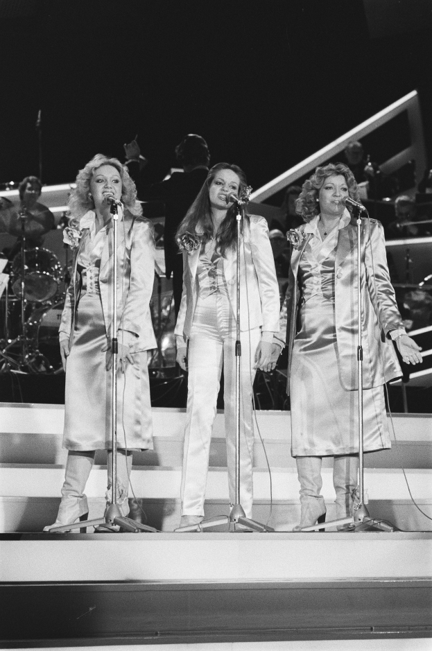 Drei Frauen singen in Mikrofone auf der Bühne mit Musikern, die hinter ihnen spielen.