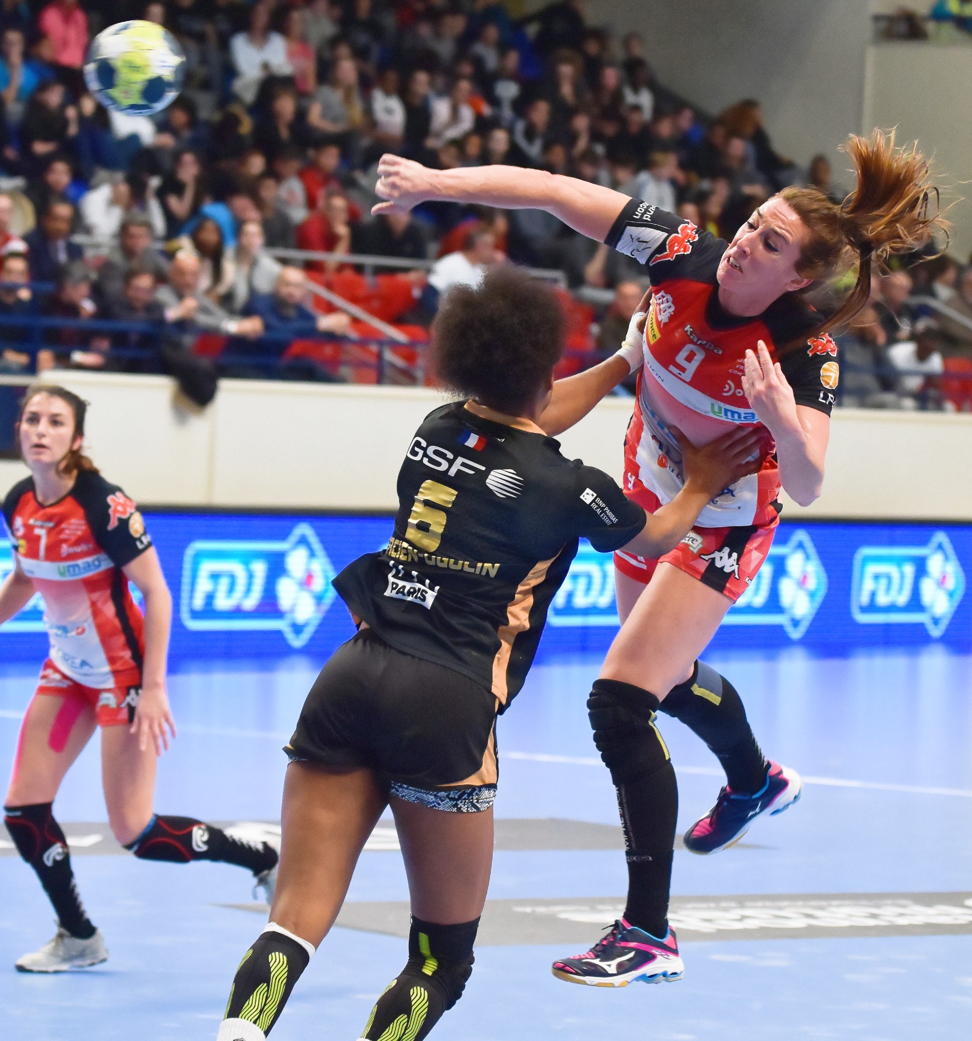 Eine Gruppe von Frauen, die auf einem Feld mit einem Ball in der Luft Handball spielen, beobachtet von Zuschauern in einer Stadionatmosphäre.