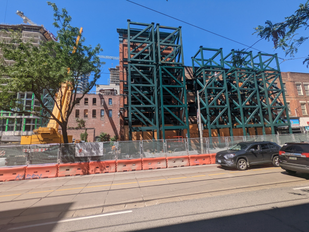 Große Metallbrücke im Bau an einer Baustelle mit parkenden Autos, einem Zaun, Bäumen, Gebäuden und Himmel im Hintergrund.