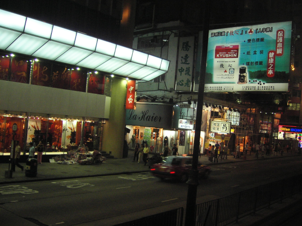 Eine nächtliche Stadtstraße mit einem vorbeifahrenden Auto vor beleuchteten Gebäuden, Schaufenstern, Fußgängern auf dem Gehweg und überirdischen Stromkabeln und Masten.