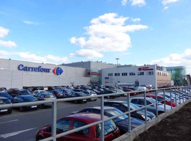 Ein großer Autohandel mit zahlreichen parkenden Autos, umgeben von Gebäuden, Straßenlaternen, Schildern, fahrenden Autos, Gittern und einem bewölkten Himmel im Hintergrund.