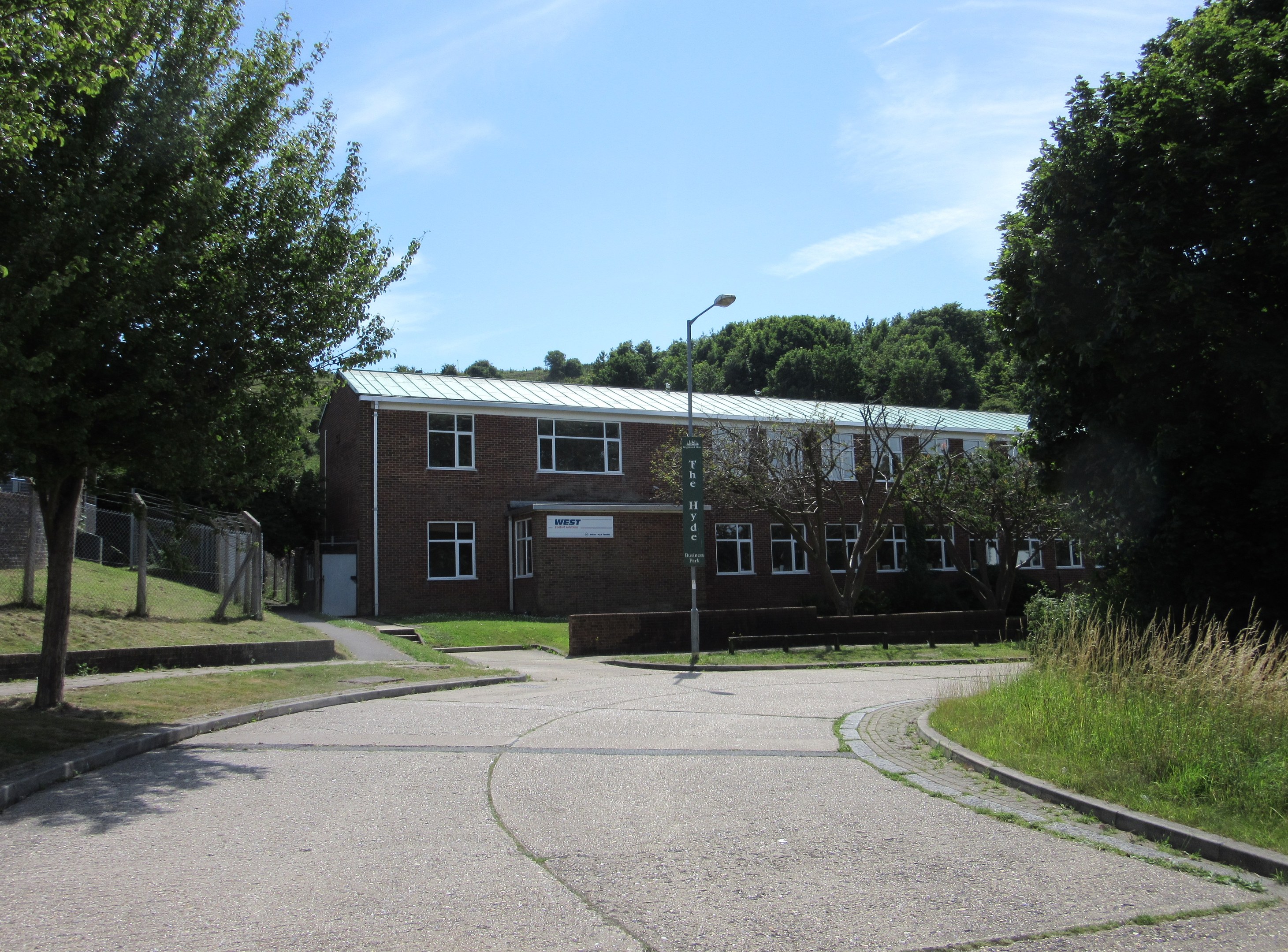 Ein Schulgebäude mit Fenstern, einer Straßenlaterne, einem Schild, einer Baumgruppe, Gras, einem Zaun und einem bewölkten Himmel, das in der Mitte eines Parks umgeben von saftigem Grün steht.
