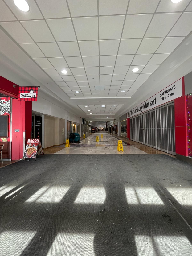 Ein hell erleuchtetes Einkaufszentrum-Innere mit einem Stuhl und einer Tafel, die links den Text "Sydbury Market - Vorher und Nachher" trägt, und verstreuten Gegenständen auf der rechten Seite, mit dem Boden darunter.