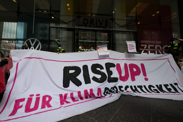 Eine Gruppe von Menschen in Masken, die eine 'Steh Auf' -Plakette und Schilder vor einem Glasgebäude bei Nacht halten.